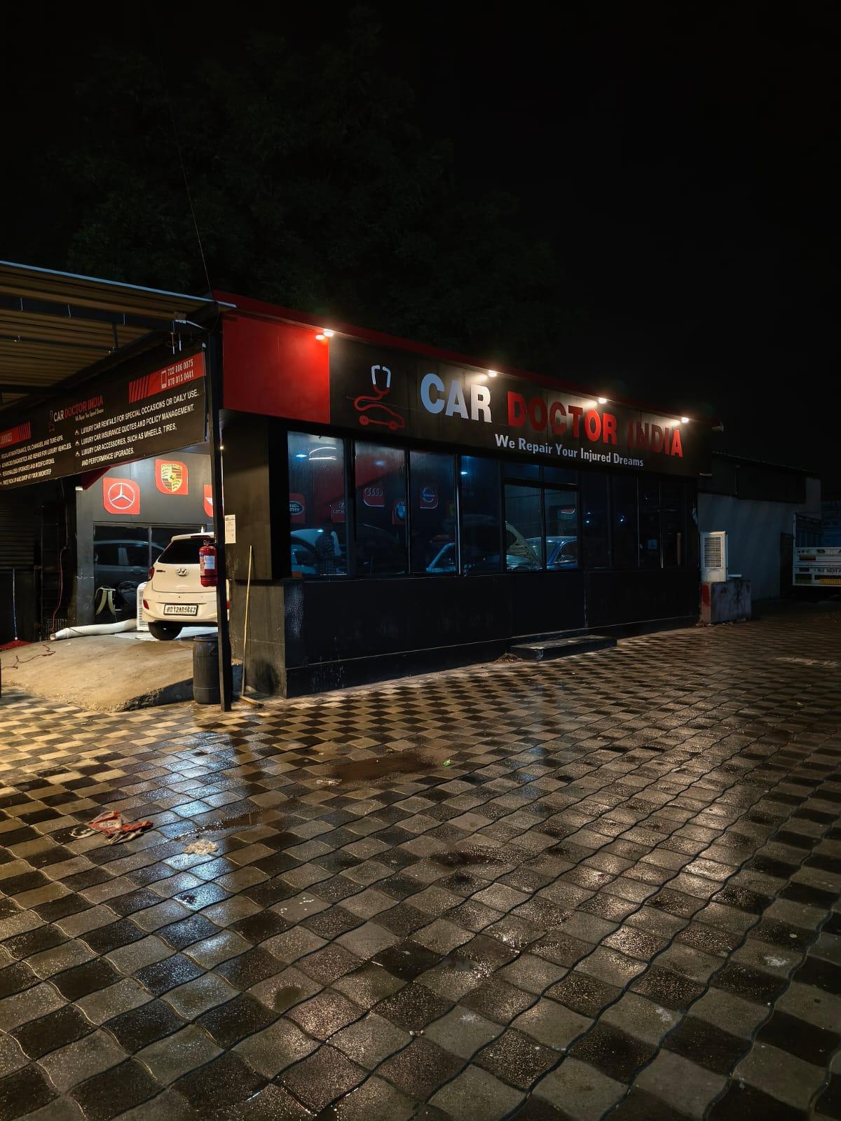 The Car Doctor India workshop on Service Road NH8 in Anand, lit up at night with its illuminated sign above the service bays